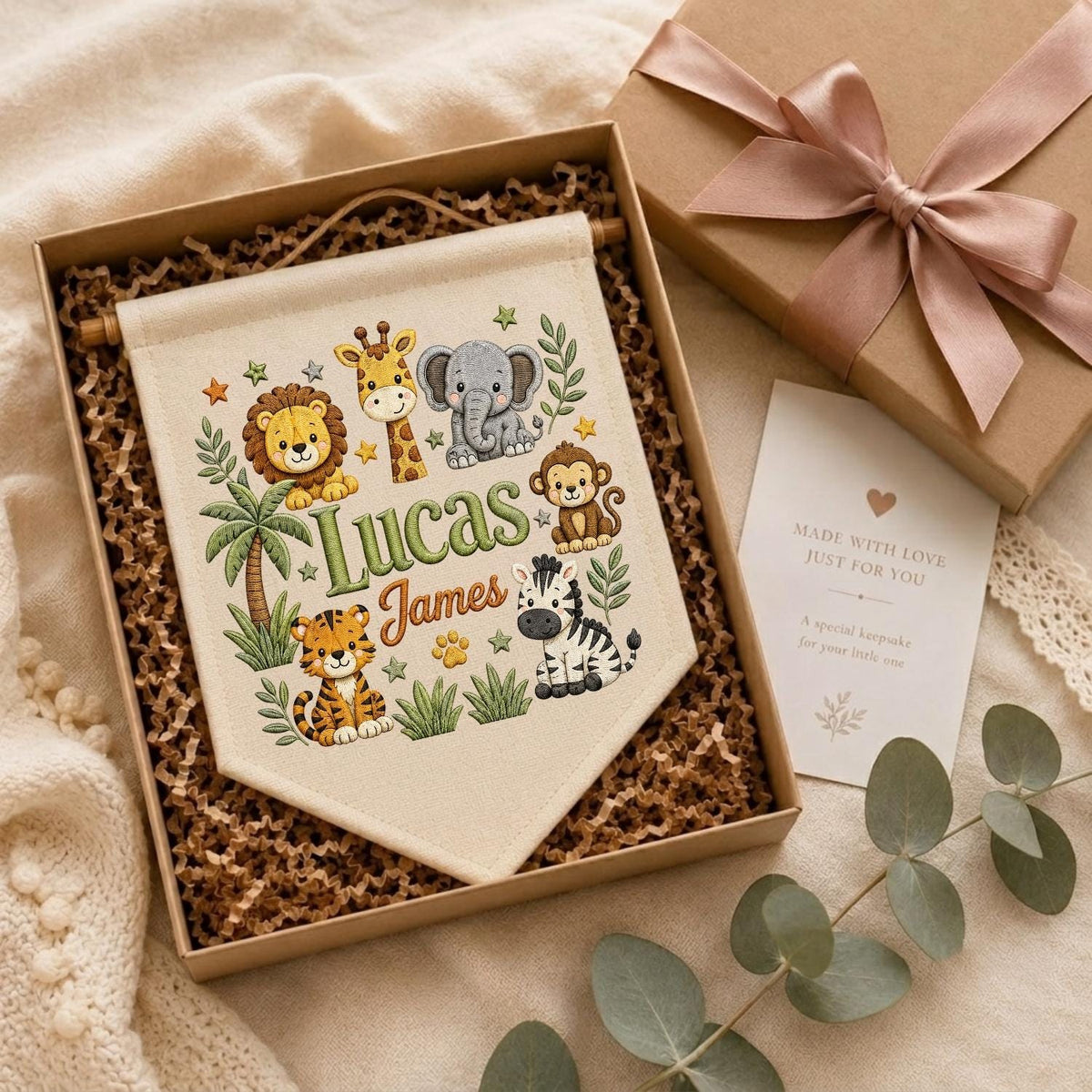 May include: A cream-colored fabric wall banner featuring a colorful illustration of a lion, giraffe, elephant, monkey, tiger, and zebra, with the names Lucas James printed in the center, presented in a gift box with a pink ribbon and card.