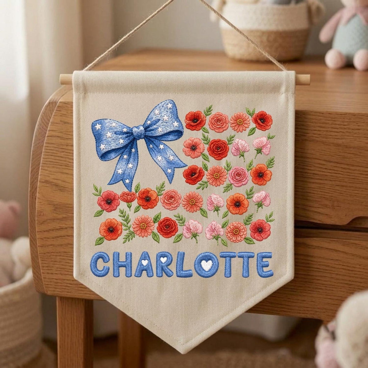 May include: A beige fabric wall hanging banner featuring an embroidered blue bow with white stars in the upper left corner. The banner displays a floral pattern of red, orange, and pink flowers with green leaves and the name CHARLOTTE in blue letters.