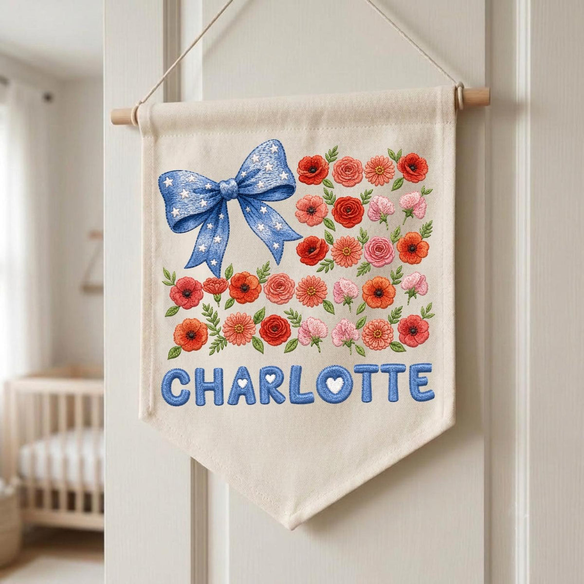 May include: A cream-colored fabric wall banner featuring a large blue bow with white stars, a collection of red, orange, and pink floral illustrations, and the name CHARLOTTE printed in bold blue letters with small white hearts inside the letters.
