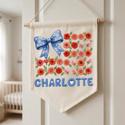 May include: A cream-colored fabric wall banner featuring a large blue bow with white stars, a collection of red, orange, and pink floral illustrations, and the name CHARLOTTE printed in bold blue letters with small white hearts inside the letters.