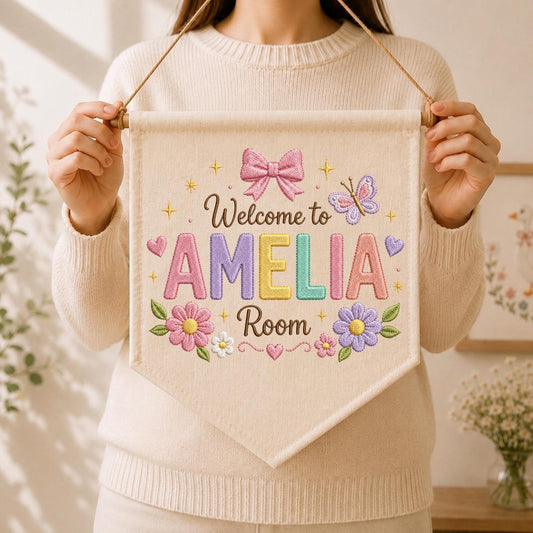 May include: A cream-colored fabric wall banner featuring embroidered text that reads Welcome to AMELIA Room. The design includes a pink bow, a butterfly, small hearts, and colorful flowers in shades of pink, yellow, and purple, all suspended by a string.