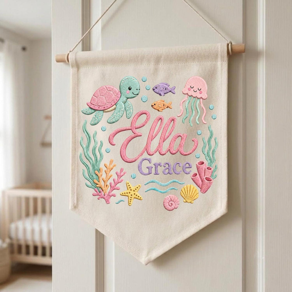 May include: A cream-colored fabric wall banner featuring embroidered sea life including a green sea turtle, pink jellyfish, starfish, and coral. The text 'Ella Grace' is embroidered in the center in pink and purple script and block lettering.