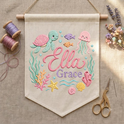 May include: A beige fabric wall hanging featuring embroidered sea life including a green turtle, pink jellyfish, orange fish, starfish, and coral. The text Ella Grace is embroidered in pink and purple script. Includes wooden dowel and string.