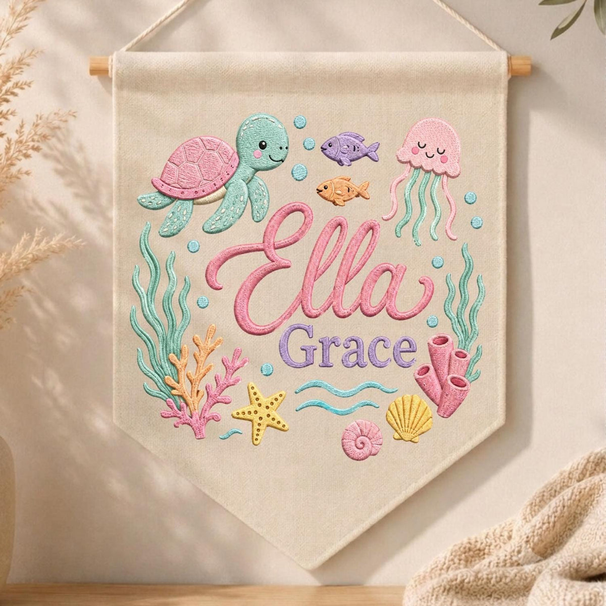 May include: A beige fabric wall hanging featuring embroidered sea life including a teal turtle, pink jellyfish, orange fish, purple fish, yellow starfish, and coral. The text Ella Grace is embroidered in the center in pink and purple script lettering.