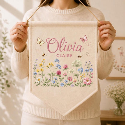 May include: A cream-colored fabric wall banner featuring the name Olivia Claire in pink script and block lettering. The design includes colorful embroidered wildflowers, two small bees, and two butterflies, suspended by a wooden dowel and string.