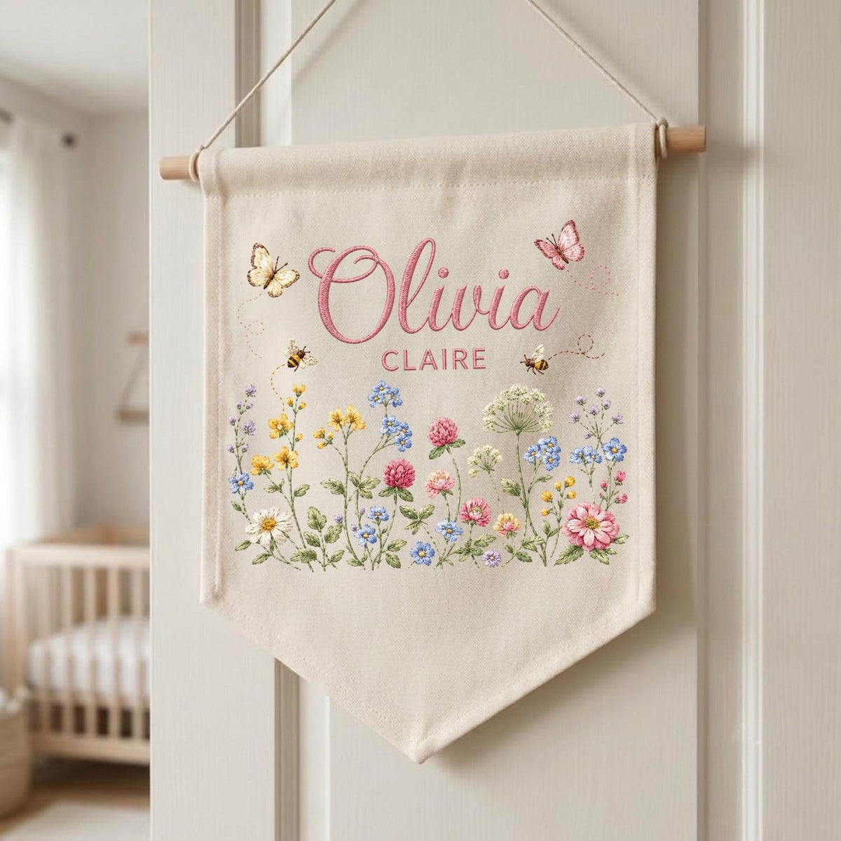 May include: A cream-colored fabric wall banner hanging from a wooden dowel with a string. The banner features the printed names Olivia and Claire in pink script and block letters, surrounded by colorful illustrations of wildflowers, bees, and butterflies.