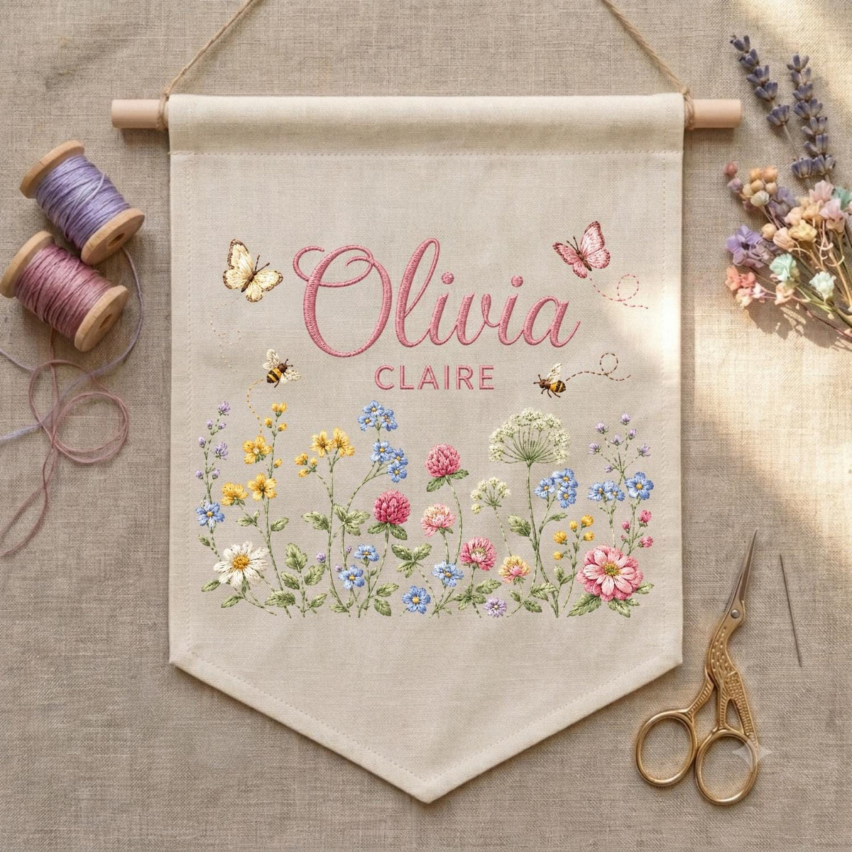 May include: A beige fabric wall banner featuring the embroidered name Olivia Claire in pink script and block letters. The design includes colorful wildflowers, two bees, and two butterflies. The banner hangs from a wooden dowel with string.