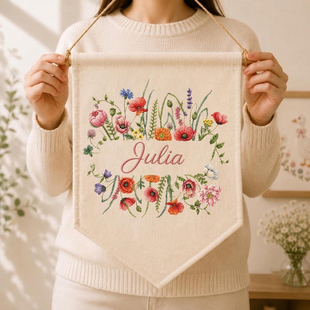 May include: A cream-colored fabric wall banner featuring an embroidered floral wreath with red, pink, orange, and purple wildflowers surrounding the name Julia in pink cursive script. The banner hangs from a wooden dowel with a thin brown cord.