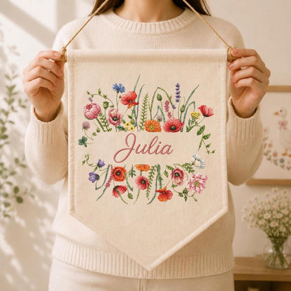 May include: A cream-colored fabric wall banner featuring an embroidered floral wreath with red, pink, orange, and purple wildflowers surrounding the name Julia in pink cursive script. The banner hangs from a wooden dowel with a thin brown cord.