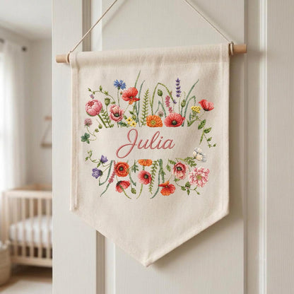 May include: A cream-colored fabric wall banner featuring an embroidered floral arrangement of red, pink, orange, blue, and green wildflowers surrounding the name Julia in pink cursive script. The banner hangs from a wooden dowel with a string.