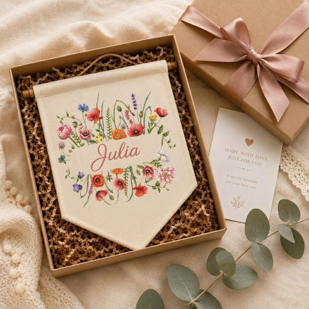 May include: A cream-colored fabric wall banner featuring an embroidered floral arrangement in red, pink, orange, blue, and green, with the name Julia in cursive script. The banner is presented in a gift box with a pink ribbon and a note.