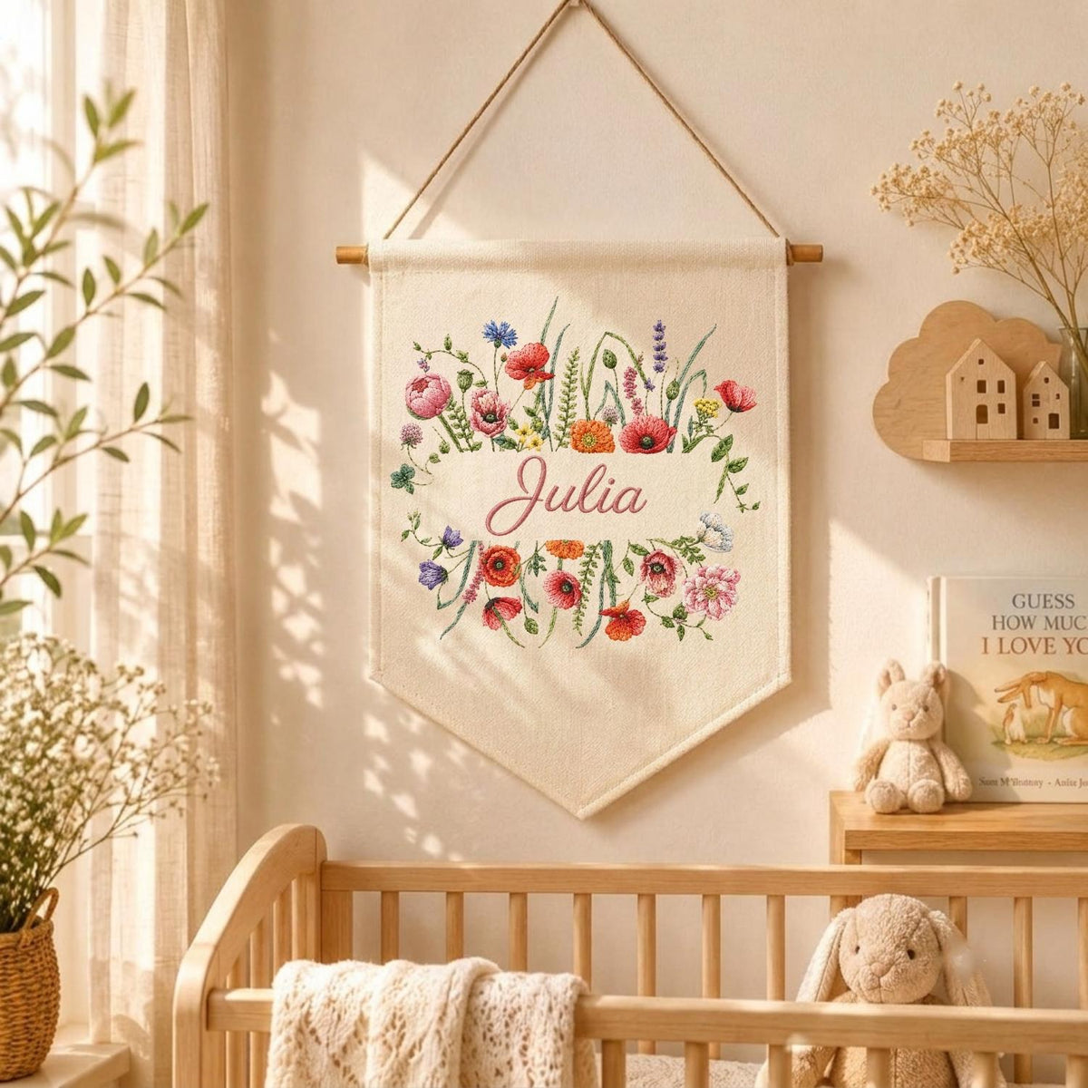 May include: A cream-colored fabric wall hanging banner featuring a colorful embroidered floral arrangement of poppies and wildflowers surrounding the name Julia in cursive script, suspended from a wooden dowel with a thin brown hanging cord.