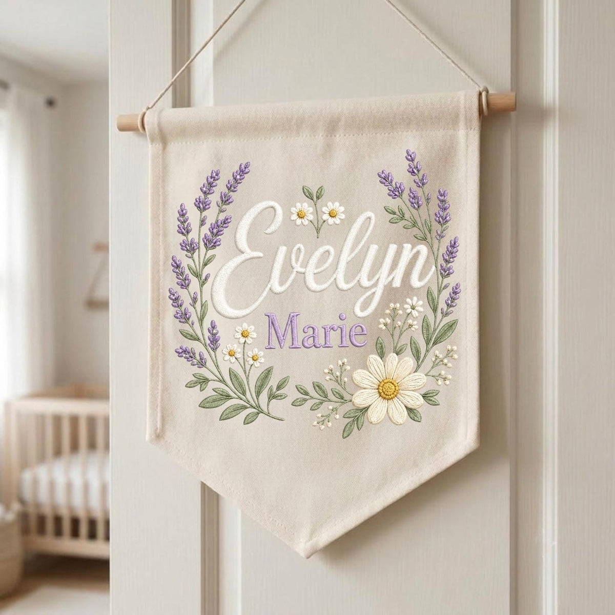 May include: A cream-colored fabric wall banner featuring the embroidered name Evelyn Marie in white and purple script. The design is framed by a floral wreath of purple lavender sprigs, small white daisies, and a large yellow-centered daisy.
