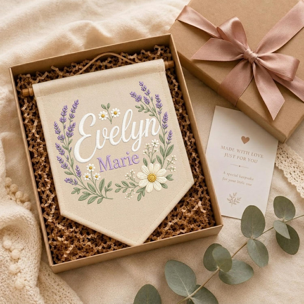 May include: A beige fabric wall banner featuring embroidered lavender sprigs and white daisies surrounding the name Evelyn Marie in white and purple script. The banner is presented in a gift box with a pink ribbon and a small card with text.