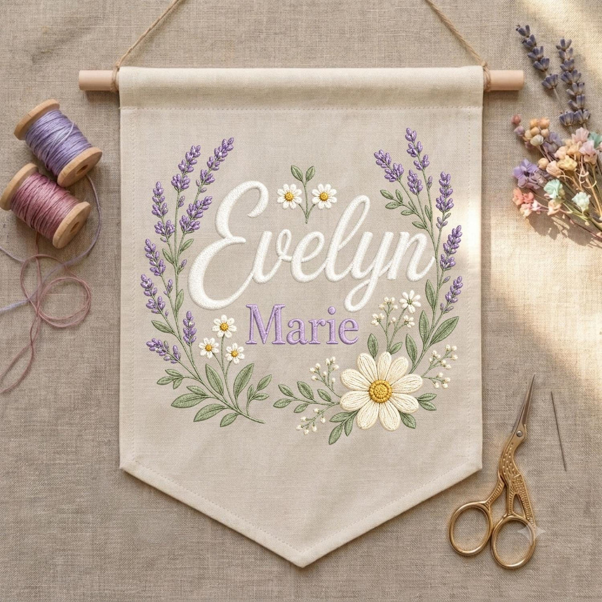 May include: A beige fabric wall banner featuring embroidered lavender sprigs and white daisies framing the name Evelyn Marie in white and purple script. The banner hangs from a wooden dowel with two spools of thread and gold scissors nearby.
