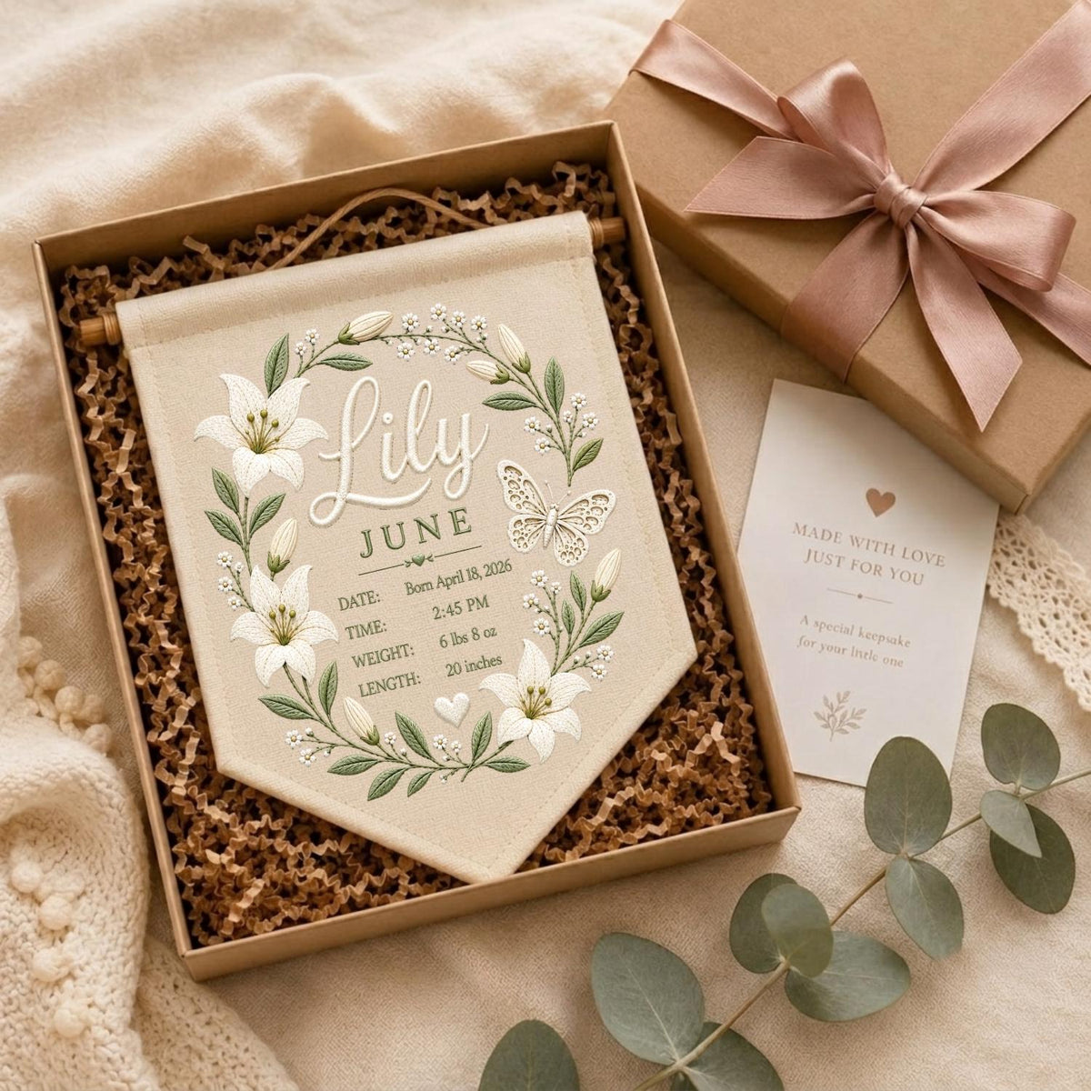 May include: A beige fabric wall banner featuring an embroidered wreath of white lilies and green leaves surrounding the name Lily, the month June, and birth details including April 18, 2026, 2:45 PM, 6 lbs 8 oz, and 20 inches, with a butterfly.