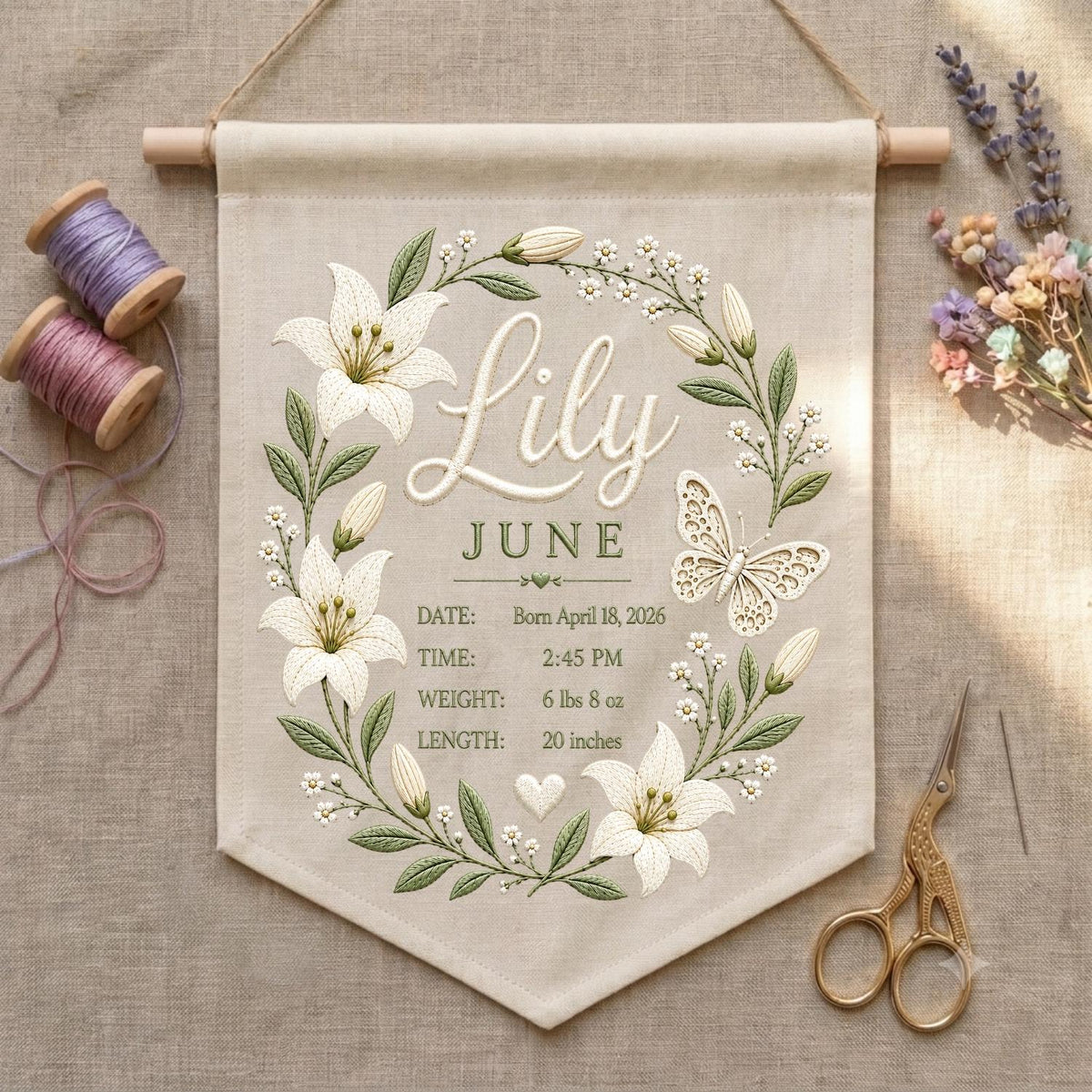 May include: A beige fabric wall hanging featuring embroidered white lily flowers, green leaves, and a butterfly. The text reads Lily, JUNE, DATE: April 18, 2026, TIME: 2:45 PM, WEIGHT: 6 lbs 8 oz, LENGTH: 20 inches. It hangs from a wooden rod with twine.