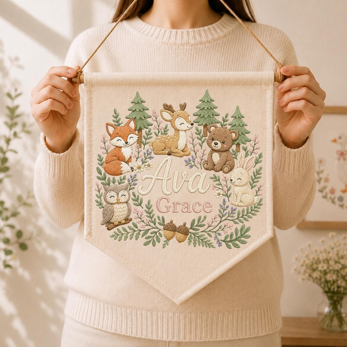 May include: A cream-colored fabric wall banner featuring an embroidered woodland scene with a fox, deer, bear, owl, and rabbit. The center displays the name Ava Grace in script and block lettering, surrounded by green pine trees, flowers, and acorns.