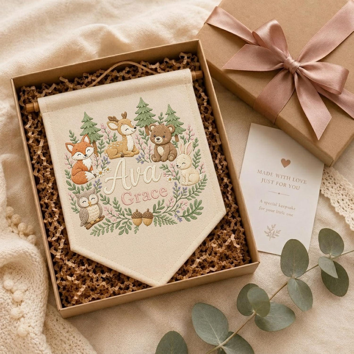 May include: A cream-colored fabric wall banner featuring an embroidered woodland scene with a fox, deer, bear, rabbit, and owl surrounded by greenery. The name Ava Grace is embroidered in the center. The banner is presented in a gift box.