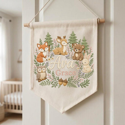 May include: A cream-colored fabric wall banner featuring a woodland theme with a fox, deer, bear, owl, and rabbit surrounded by green trees and pink flowers. The name Ava Grace is embroidered in the center in a script and block font.