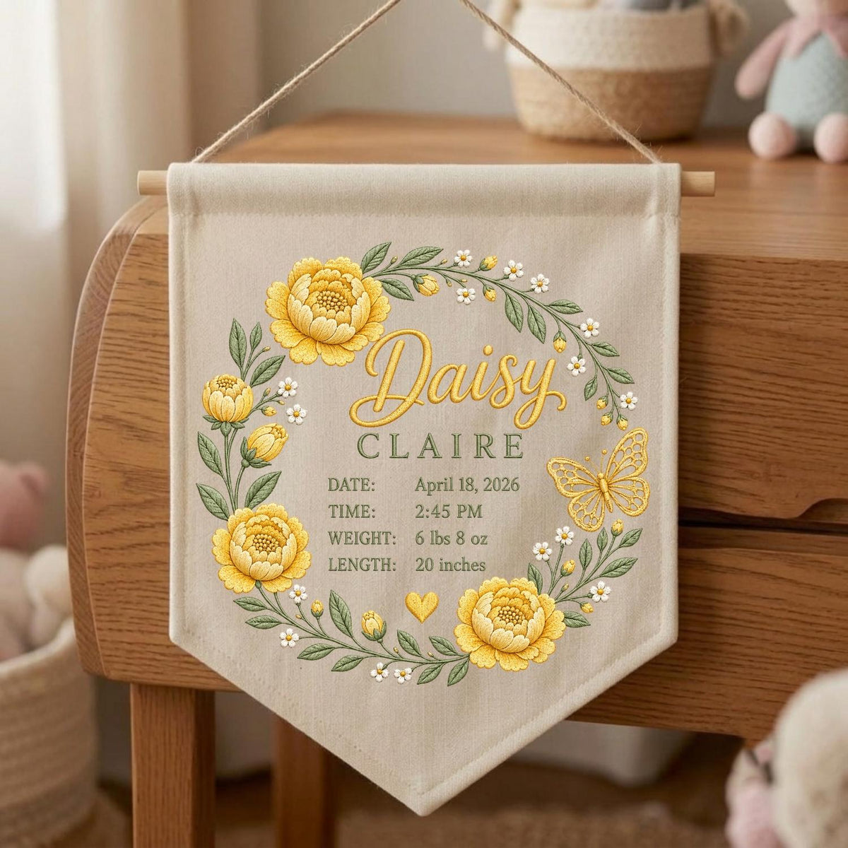May include: A beige fabric wall banner featuring a yellow floral wreath with a butterfly. The text reads: Daisy CLAIRE, DATE: April 18, 2026, TIME: 2:45 PM, WEIGHT: 6 lbs 8 oz, LENGTH: 20 inches. The banner hangs from a wooden dowel with a string.