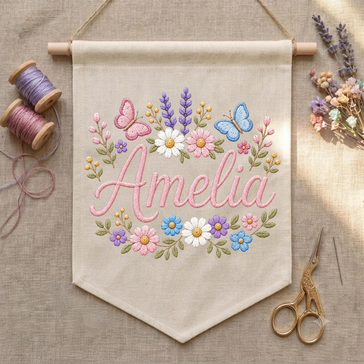May include: A beige fabric wall banner featuring the embroidered name Amelia in pink script, surrounded by a floral wreath of pink, white, blue, and purple flowers, green leaves, and two butterflies, suspended from a wooden dowel with string.