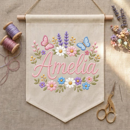 May include: A beige fabric wall banner featuring the embroidered name Amelia in pink script, surrounded by a floral wreath of pink, white, blue, and purple flowers, green leaves, and two butterflies, suspended from a wooden dowel with string.