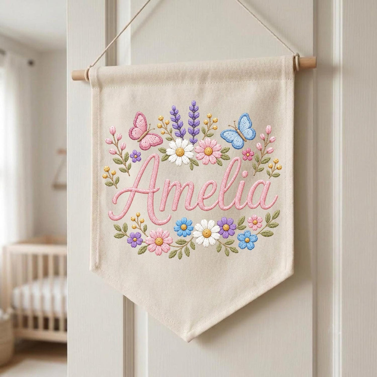 May include: A beige fabric wall banner featuring the embroidered name Amelia in pink script, surrounded by a colorful floral wreath with daisies, lavender, and small blossoms, accented by two embroidered butterflies in pink and blue hues.