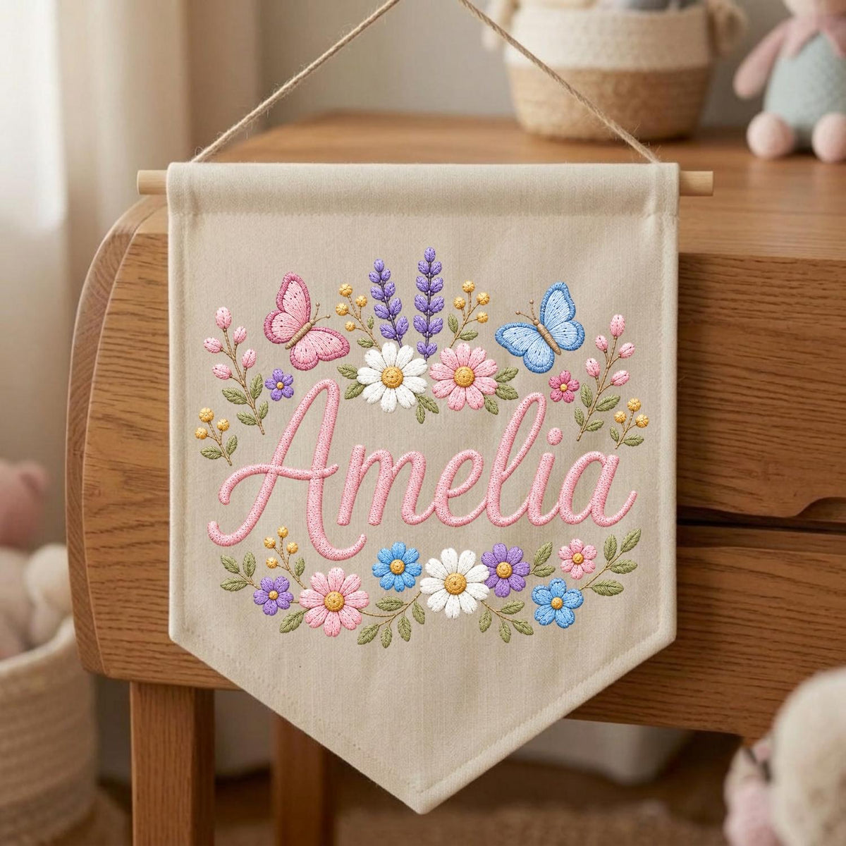 May include: A beige fabric wall banner featuring the embroidered name Amelia in pink script. The design includes a colorful floral wreath with pink, white, blue, and purple flowers, green leaves, and two butterflies in pink and blue shades.