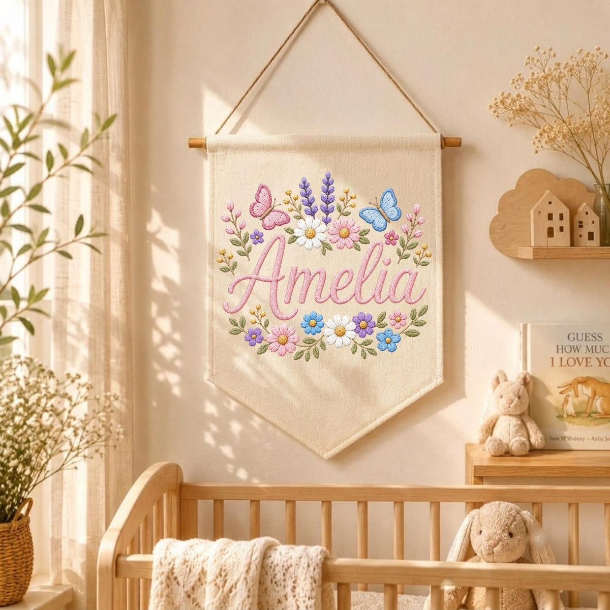 May include: A cream-colored fabric wall hanging banner featuring the name Amelia embroidered in pink script, surrounded by a colorful floral wreath with pink, blue, and white flowers, green leaves, and two butterflies, suspended from a wooden rod.