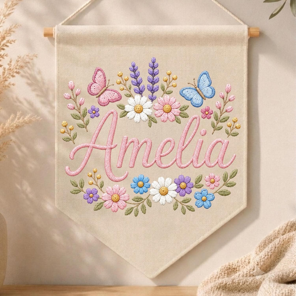 May include: A beige fabric wall banner featuring the embroidered name Amelia in pink script. The design includes colorful floral motifs with daisies, lavender, and small blossoms, accented by a pink butterfly on the left and a blue butterfly on the right.