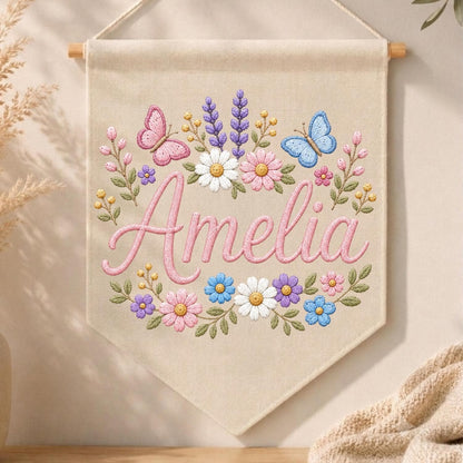May include: A beige fabric wall banner featuring the embroidered name Amelia in pink script. The design includes colorful floral motifs with daisies, lavender, and small blossoms, accented by a pink butterfly on the left and a blue butterfly on the right.