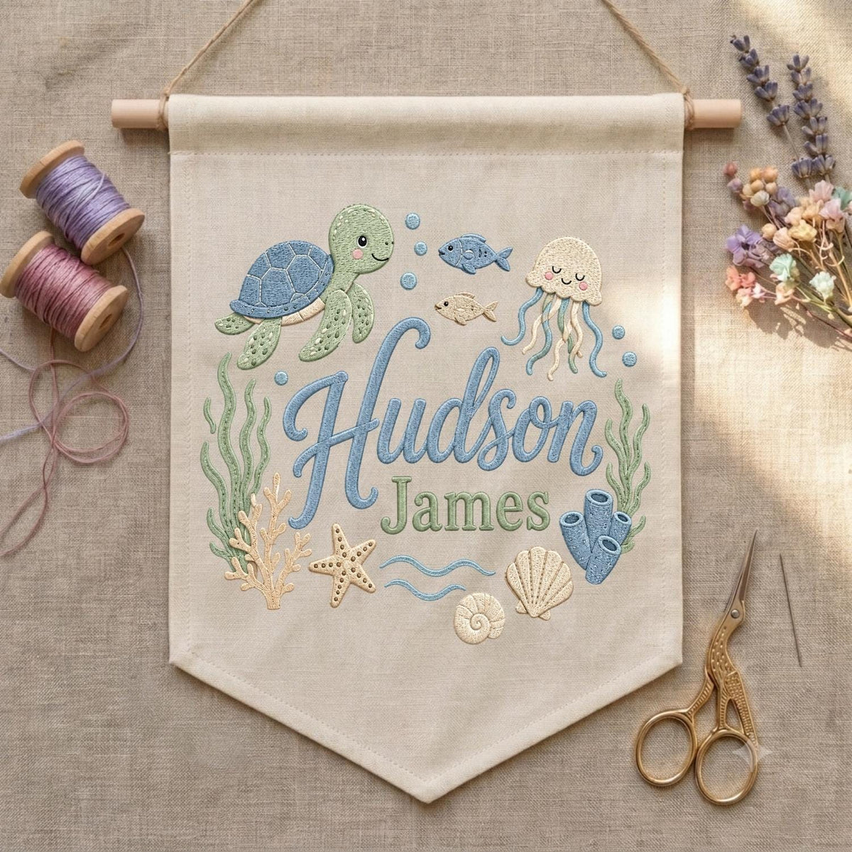 May include: A beige fabric wall banner featuring embroidered ocean life including a green sea turtle, a jellyfish, a starfish, seashells, and seaweed. The text "Hudson James" is embroidered in blue and green script in the center.