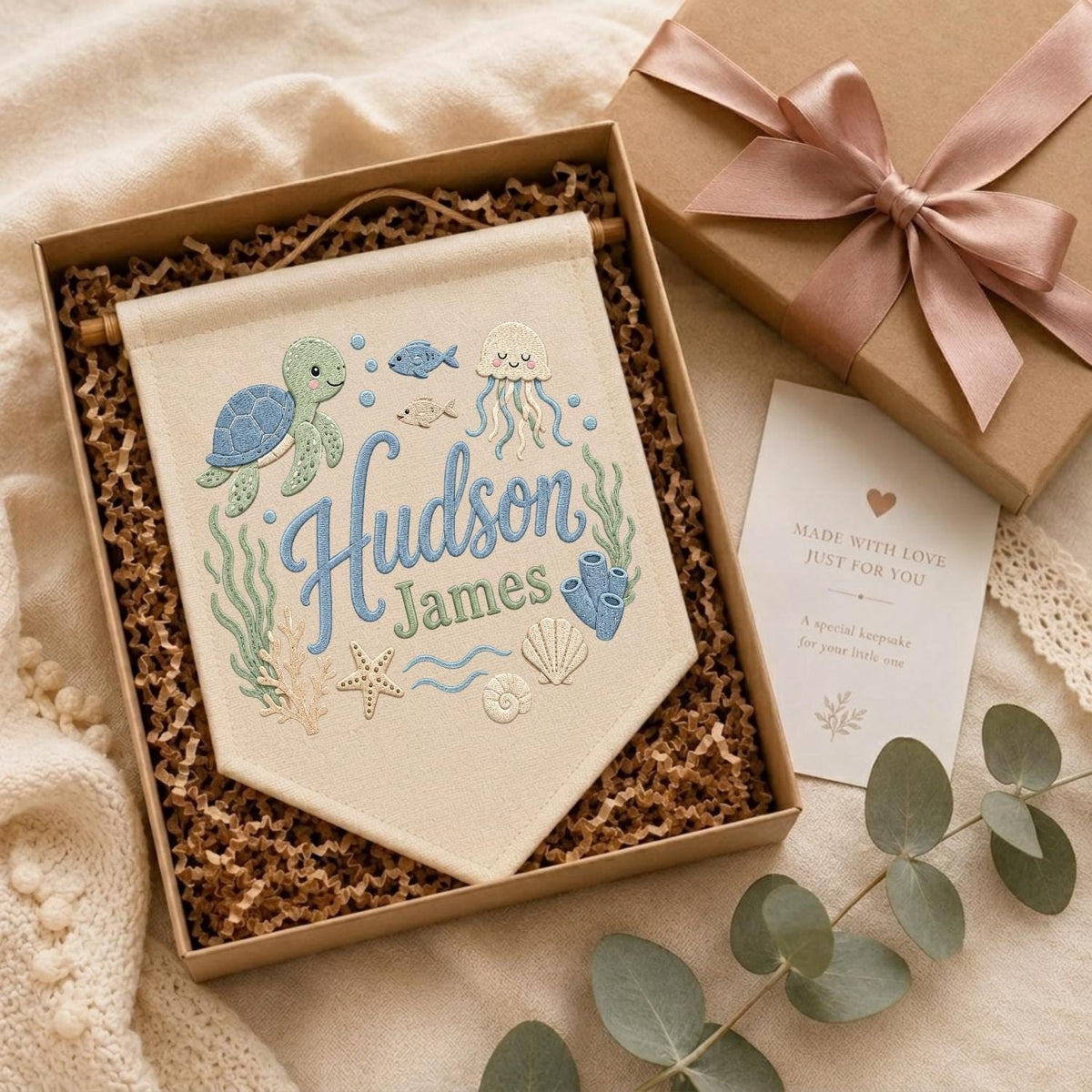 May include: A cream-colored fabric wall banner featuring embroidered sea life including a green turtle, jellyfish, starfish, and shells. The text reads Hudson James in blue and green script. The banner is inside a gift box with a pink ribbon.