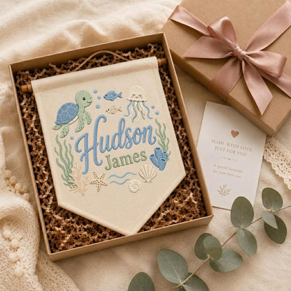 May include: A cream-colored fabric wall banner featuring embroidered sea life including a green turtle, jellyfish, starfish, and shells. The text reads Hudson James in blue and green script. The banner is inside a gift box with a pink ribbon.