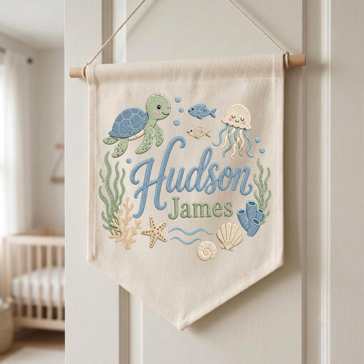 May include: A cream-colored fabric wall banner featuring embroidered ocean-themed designs including a green sea turtle, a jellyfish, fish, coral, a starfish, and seashells. The text "Hudson James" is embroidered in blue and green script.
