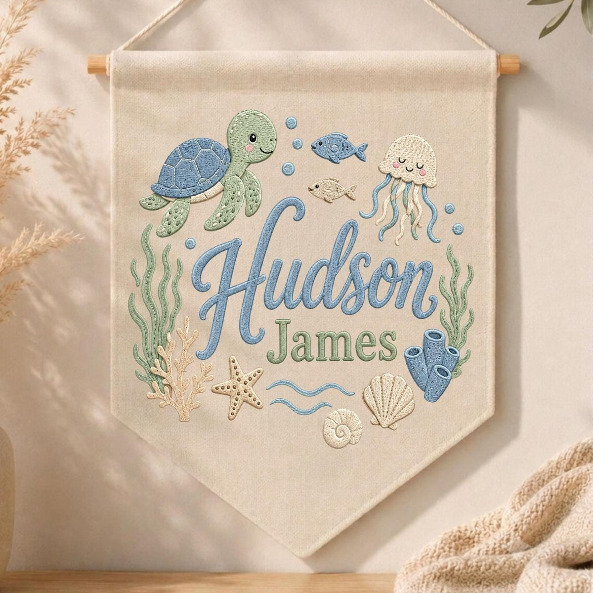 May include: A cream-colored fabric wall banner featuring embroidered ocean life including a blue sea turtle, a jellyfish, a starfish, coral, and seaweed. The text "Hudson James" is embroidered in the center in blue and green script lettering.