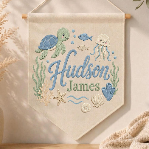 May include: A cream-colored fabric wall banner featuring embroidered ocean life including a blue sea turtle, a jellyfish, a starfish, coral, and seaweed. The text "Hudson James" is embroidered in the center in blue and green script lettering.