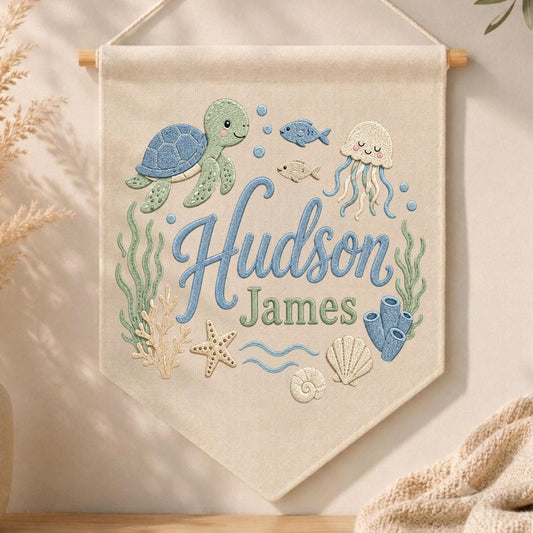 May include: A cream-colored fabric wall banner featuring embroidered ocean life including a blue sea turtle, a jellyfish, a starfish, coral, and seaweed. The text "Hudson James" is embroidered in the center in blue and green script lettering.