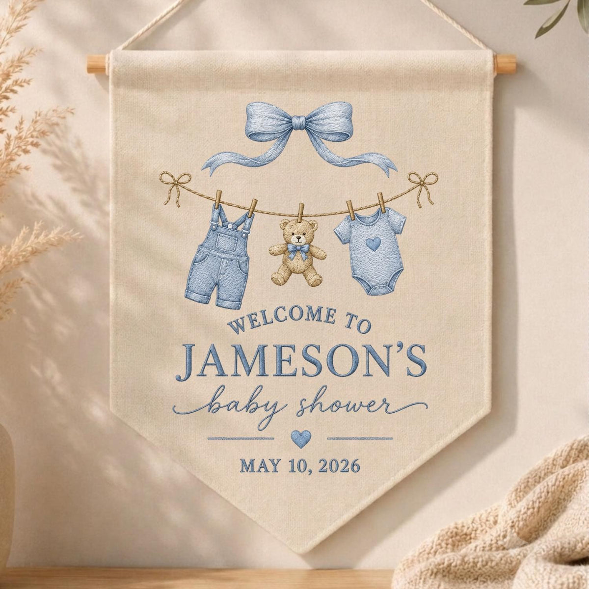 May include: A beige fabric wall banner featuring a blue bow, a teddy bear, and baby clothes hanging on a line. The text reads: WELCOME TO JAMESON'S baby shower MAY 10, 2026. The banner is suspended from a wooden rod with a white string for hanging.