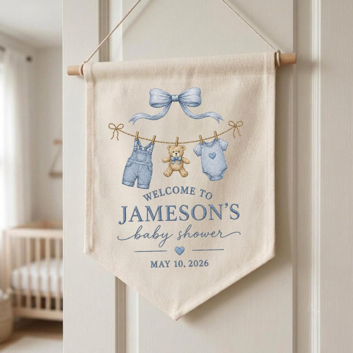 May include: A cream-colored fabric wall banner featuring a blue bow, a teddy bear, and baby clothes hanging on a line. The text reads: WELCOME TO JAMESON'S baby shower MAY 10, 2026. The banner is suspended from a wooden dowel with a string.