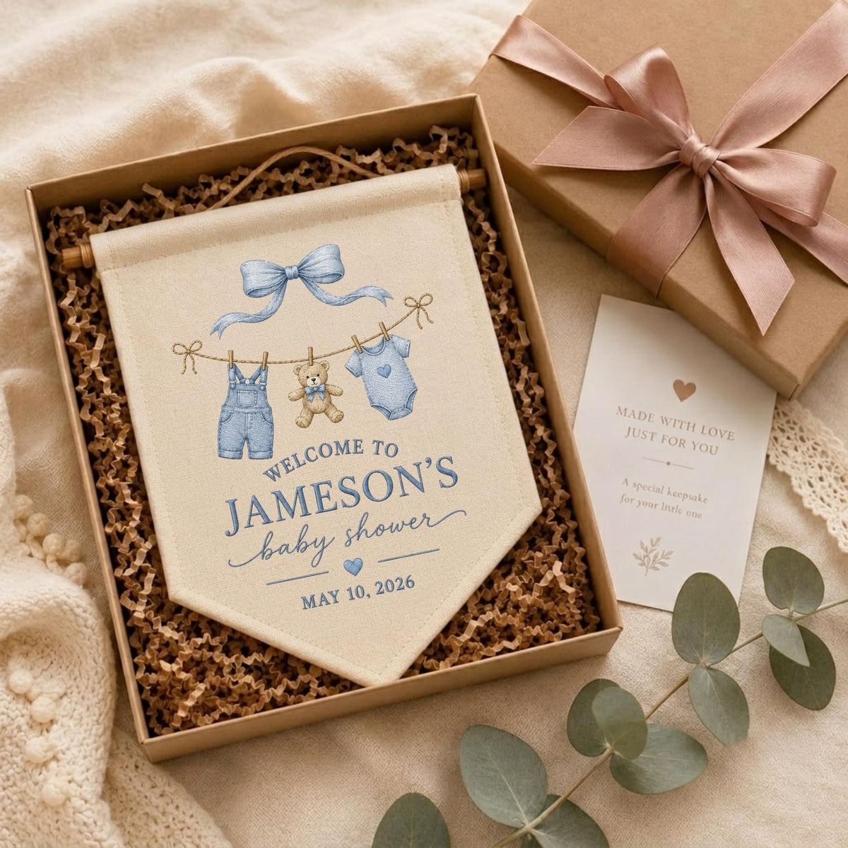 May include: A cream-colored fabric banner featuring a blue bow, a teddy bear, and baby clothes hanging on a line. The text reads WELCOME TO JAMESON'S baby shower MAY 10, 2026. The banner is inside a gift box with a pink ribbon and a small card.