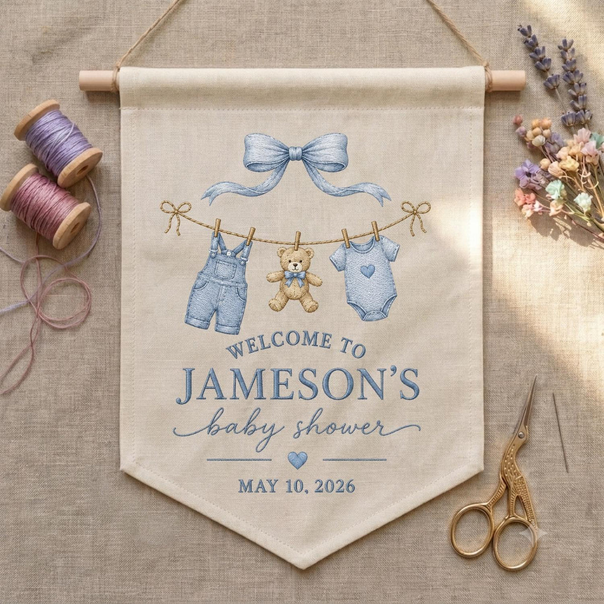 May include: A beige fabric wall banner featuring a blue bow, a clothesline with blue overalls, a teddy bear, and a blue onesie. The text reads: WELCOME TO JAMESON'S baby shower MAY 10, 2026. The banner hangs from a wooden dowel with a string.