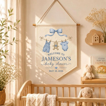 May include: A cream-colored fabric wall banner featuring a blue bow, a teddy bear, and baby clothes hanging on a line. The text reads: WELCOME TO JAMESON'S baby shower MAY 10, 2026. The banner is suspended from a wooden rod with a hanging cord.