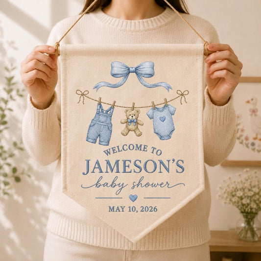 May include: A cream-colored fabric banner featuring a blue bow, a clothesline with denim overalls, a teddy bear, and a blue onesie. The text reads: WELCOME TO JAMESON'S baby shower MAY 10, 2026. The banner hangs from a wooden dowel with a string.