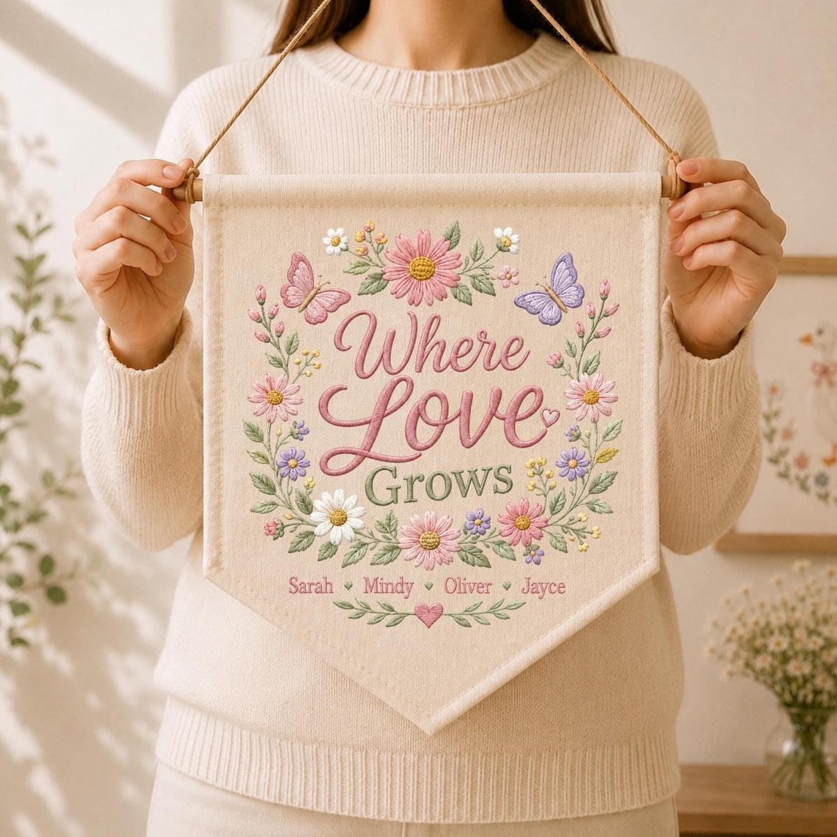 May include: A cream-colored fabric wall banner featuring embroidered floral wreaths in pink, purple, and white with two butterflies. The text reads Where Love Grows, followed by the names Sarah, Mindy, Oliver, and Jayce, suspended by a wooden rod.