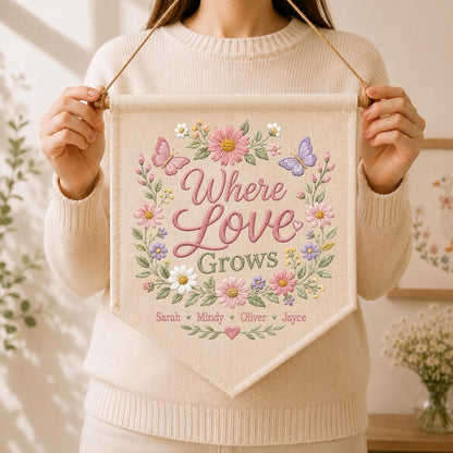 May include: A cream-colored fabric wall banner featuring embroidered floral wreaths in pink, purple, and white with two butterflies. The text reads Where Love Grows, followed by the names Sarah, Mindy, Oliver, and Jayce, suspended by a wooden rod.
