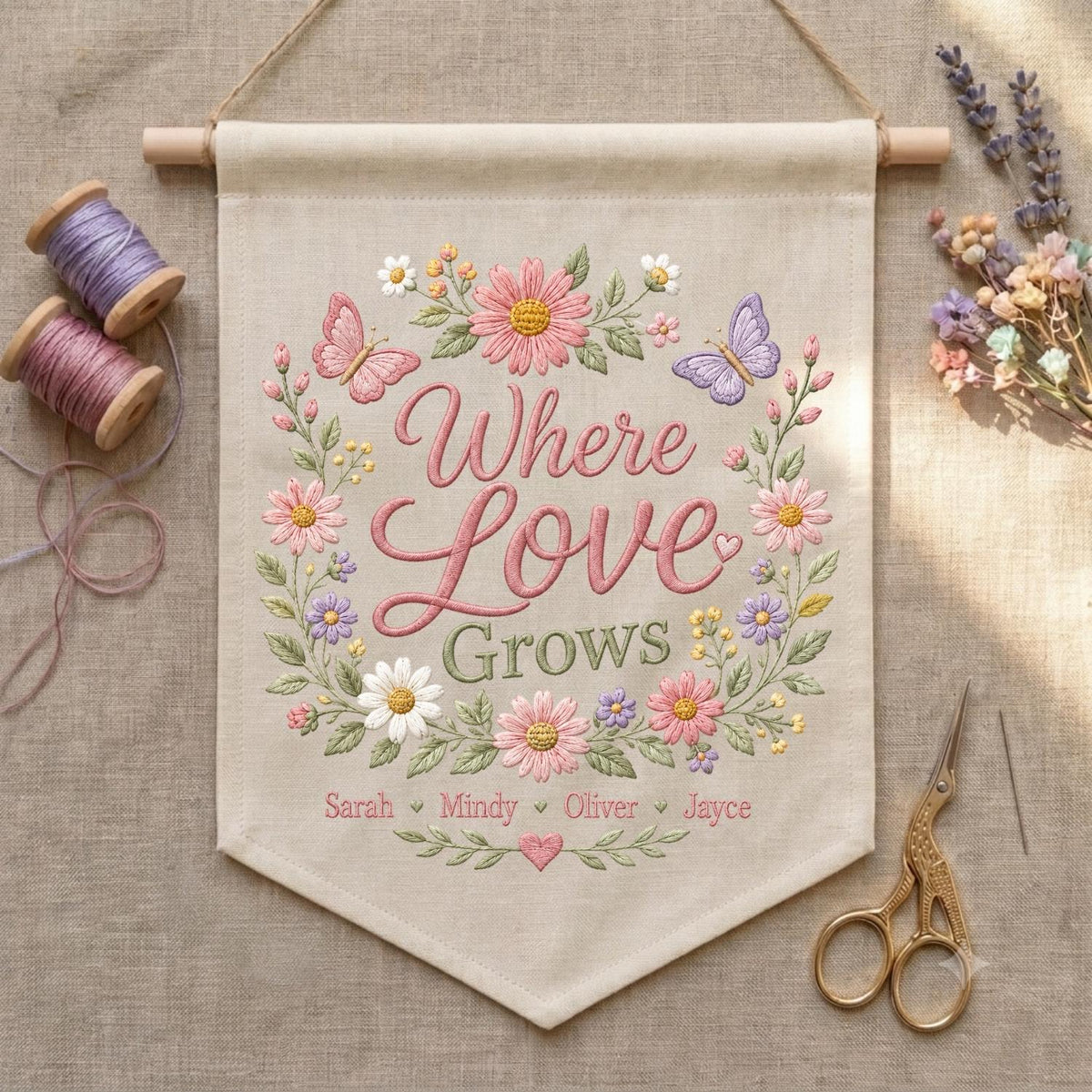 May include: A beige fabric wall hanging featuring embroidered floral wreaths and butterflies surrounding the text Where Love Grows. Below the text are the names Sarah, Mindy, Oliver, and Jayce in pink thread, with a small heart icon.