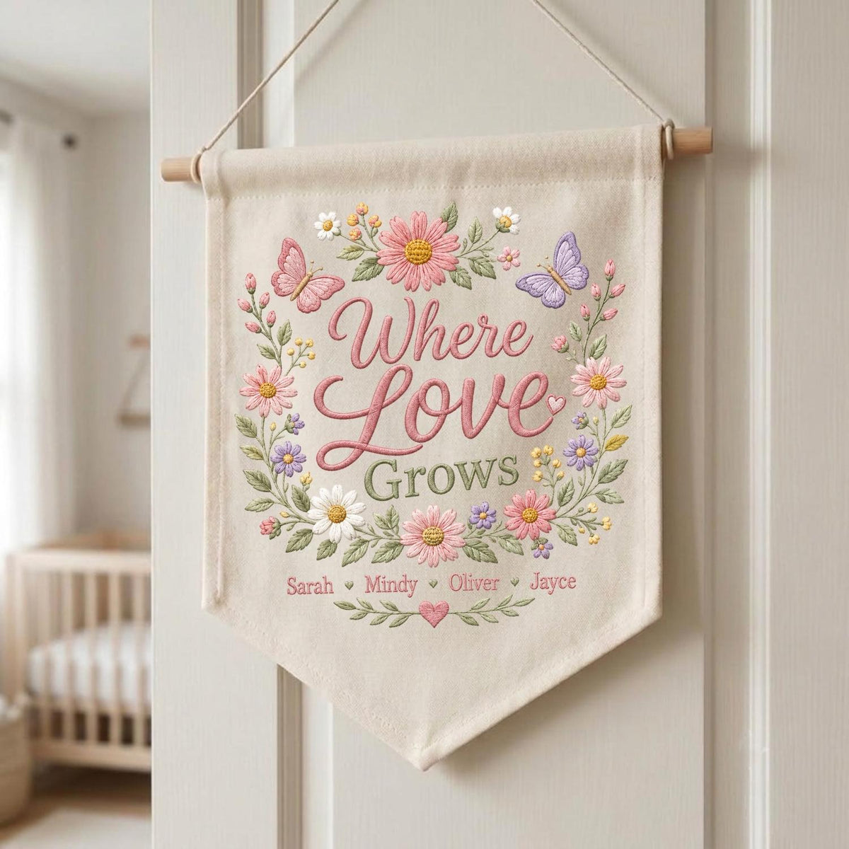 May include: A cream-colored fabric wall banner hanging from a wooden dowel with string. It features embroidered pink, white, and purple flowers, green leaves, and two butterflies. The text reads Where Love Grows, with the names Sarah, Mindy, Oliver, and Jayce.