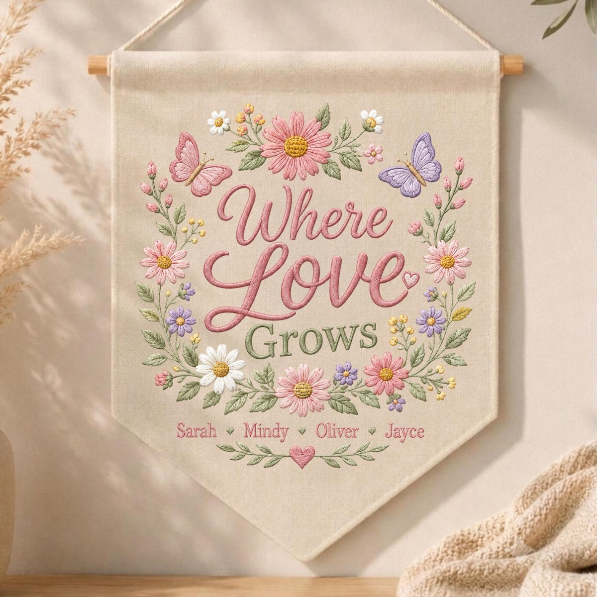 May include: A beige fabric wall banner featuring embroidered floral wreaths in pink, purple, and white with two butterflies. The text reads Where Love Grows, followed by the names Sarah, Mindy, Oliver, and Jayce, separated by small hearts.